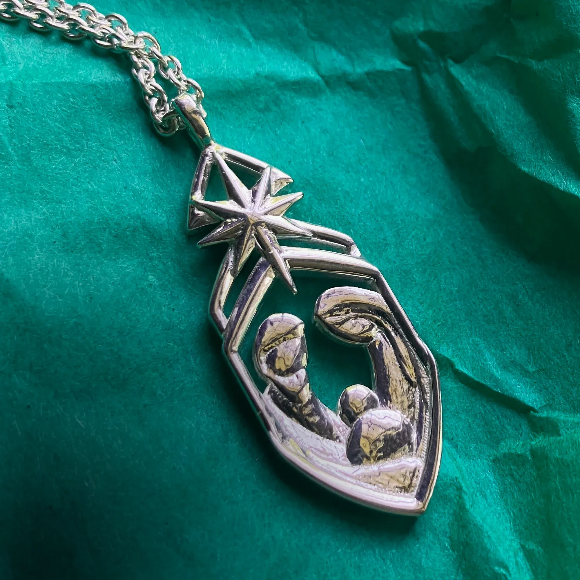 Silver nativity pendant, a star above a stylized holy family, lying on green tissue paper.